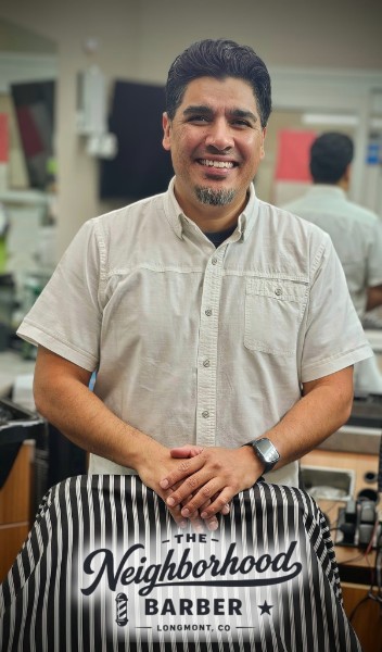 Jose, the owner of The Neighborhood Barber, Longmont standing behind a barber chair while smiling.